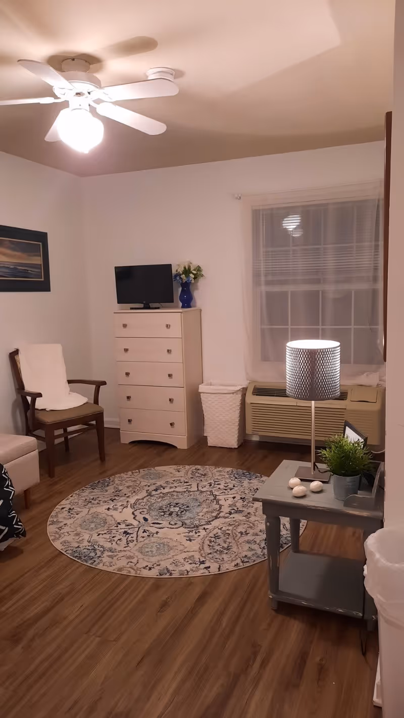 Small furnished living room with a round patterned rug, dresser with TV, chair, side table with lamp and a window air conditioner.
