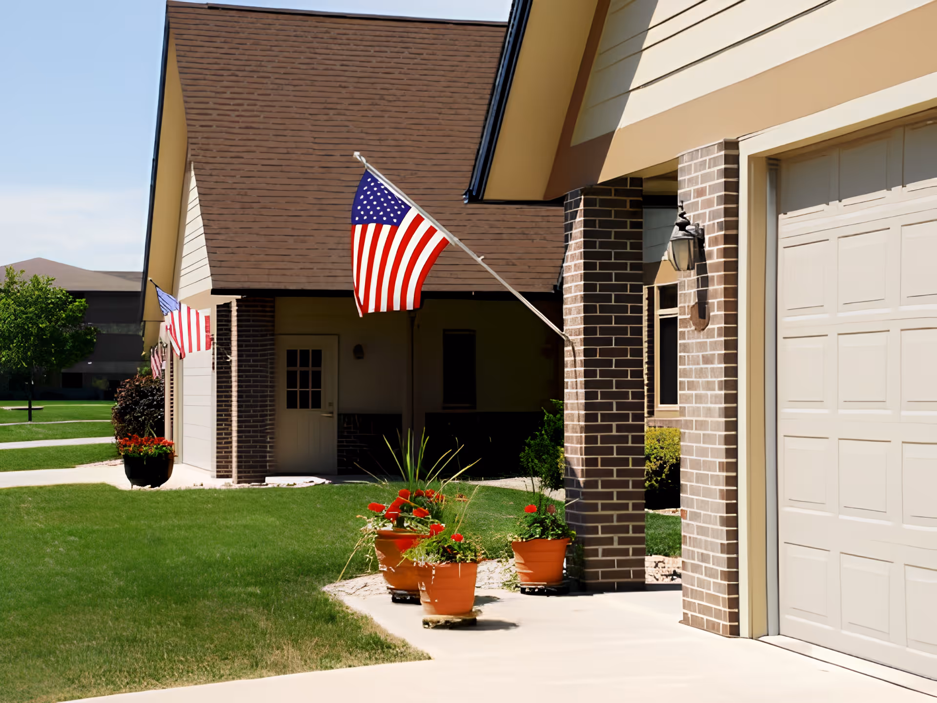 Exterior view of a senior living facility with brick walls and beige garage doors. Several American flags are mounted on the building, and there are potted plants with red flowers along the walkway. The lawn is green and well-maintained under a clear blue sky.