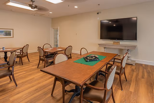 Interior communal dining/activity room with multiple wooden tables and chairs, a wall-mounted TV, and wood flooring.