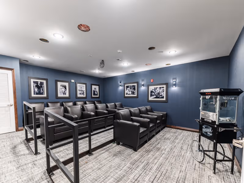 A small movie theater room with two rows of black leather armchairs separated by a black metal railing. The walls are painted dark blue and decorated with black and white framed photographs. A vintage-style popcorn machine on wheels is positioned near the entrance door. The ceiling has several recessed lights and some exposed wiring or fixtures.
