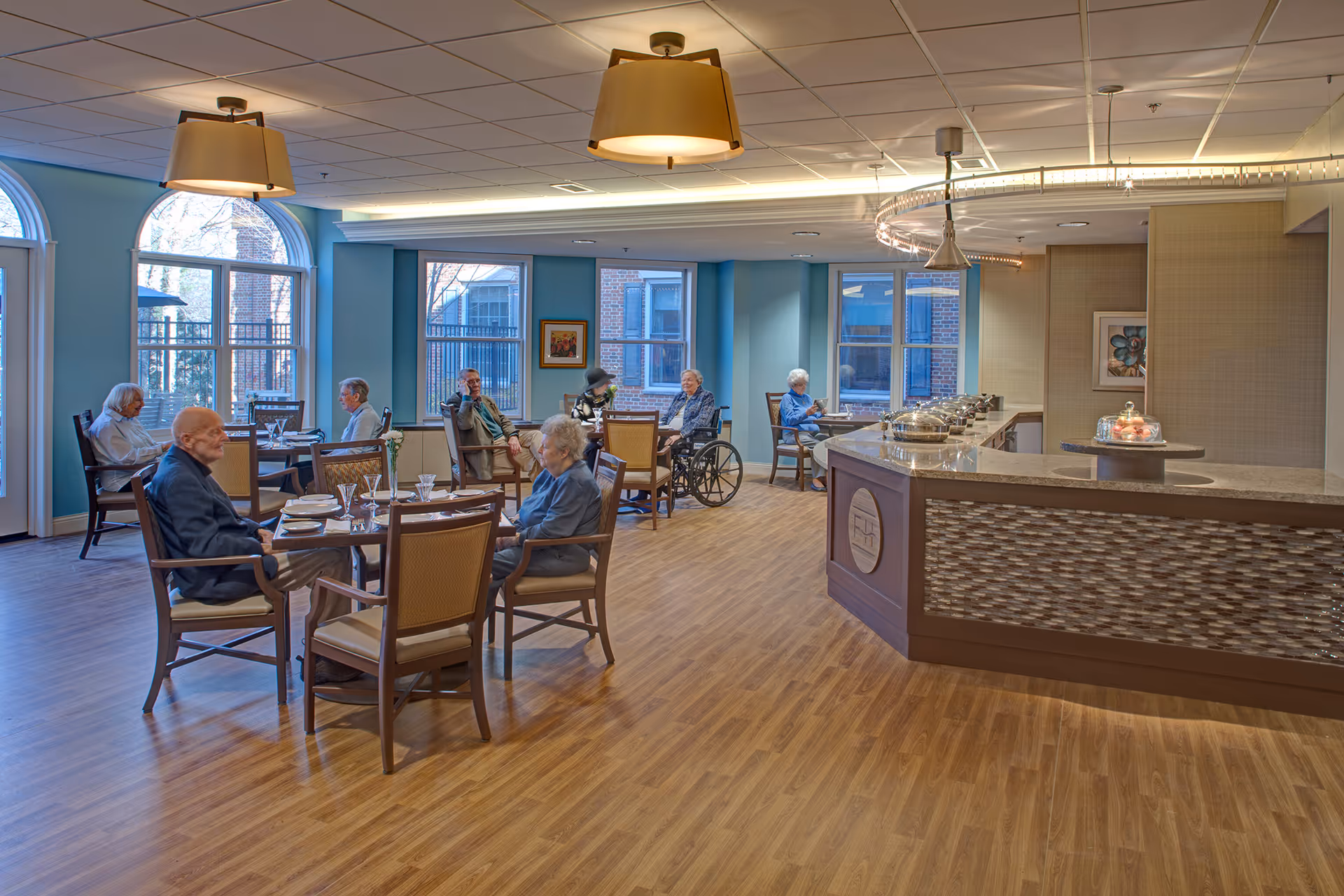 A dining area in a senior living facility with several elderly residents seated at tables. The room has large windows with blue walls, wooden flooring, and modern light fixtures. There is a buffet counter on the right side with covered dishes and a cake stand.