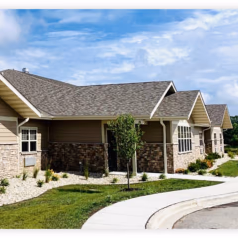 Exterior view of a single-story assisted living facility building with beige siding and stone accents, multiple windows, a shingled roof, a small tree, and landscaped greenery along a curved sidewalk under a partly cloudy sky.