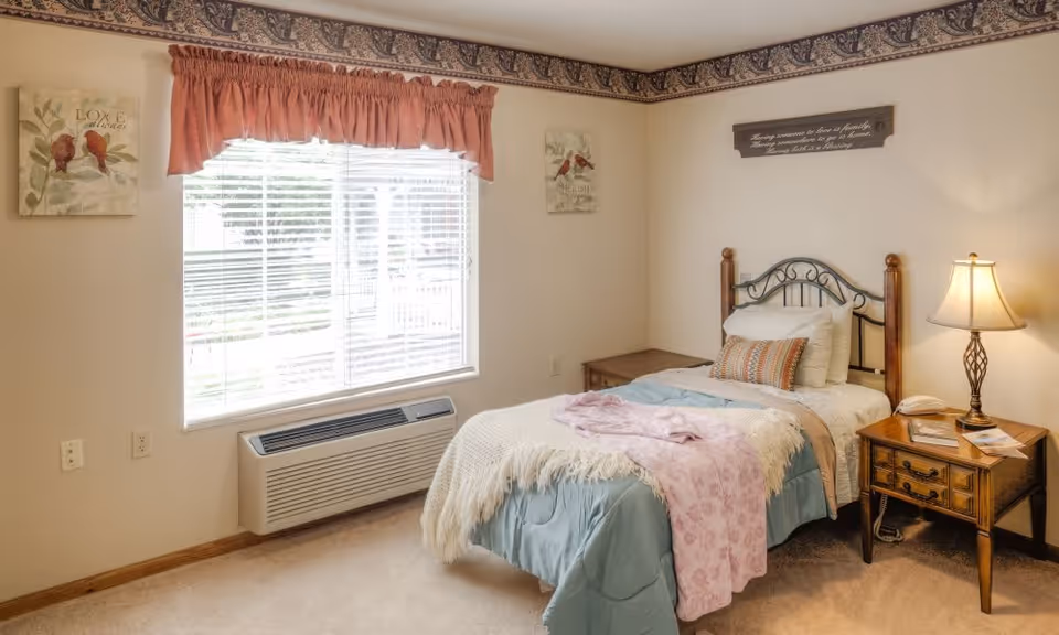 A cozy bedroom with a single bed dressed in blue and white bedding with a pink throw blanket. The bed has a wooden and metal headboard. Next to the bed is a wooden nightstand with a lamp, a telephone, and some papers. The room has a window with white blinds and a pink valance, two framed bird-themed wall art pieces, and a decorative border near the ceiling.