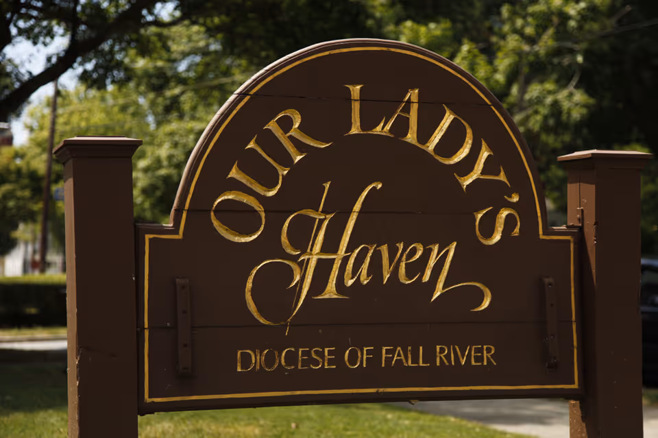 A brown wooden sign with gold lettering that reads 'Our Lady's Haven Diocese of Fall River' outdoors with trees and grass in the background.