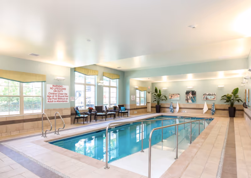Bright indoor swimming pool area with handrails, seating along windows, potted plants, and pool safety signage.