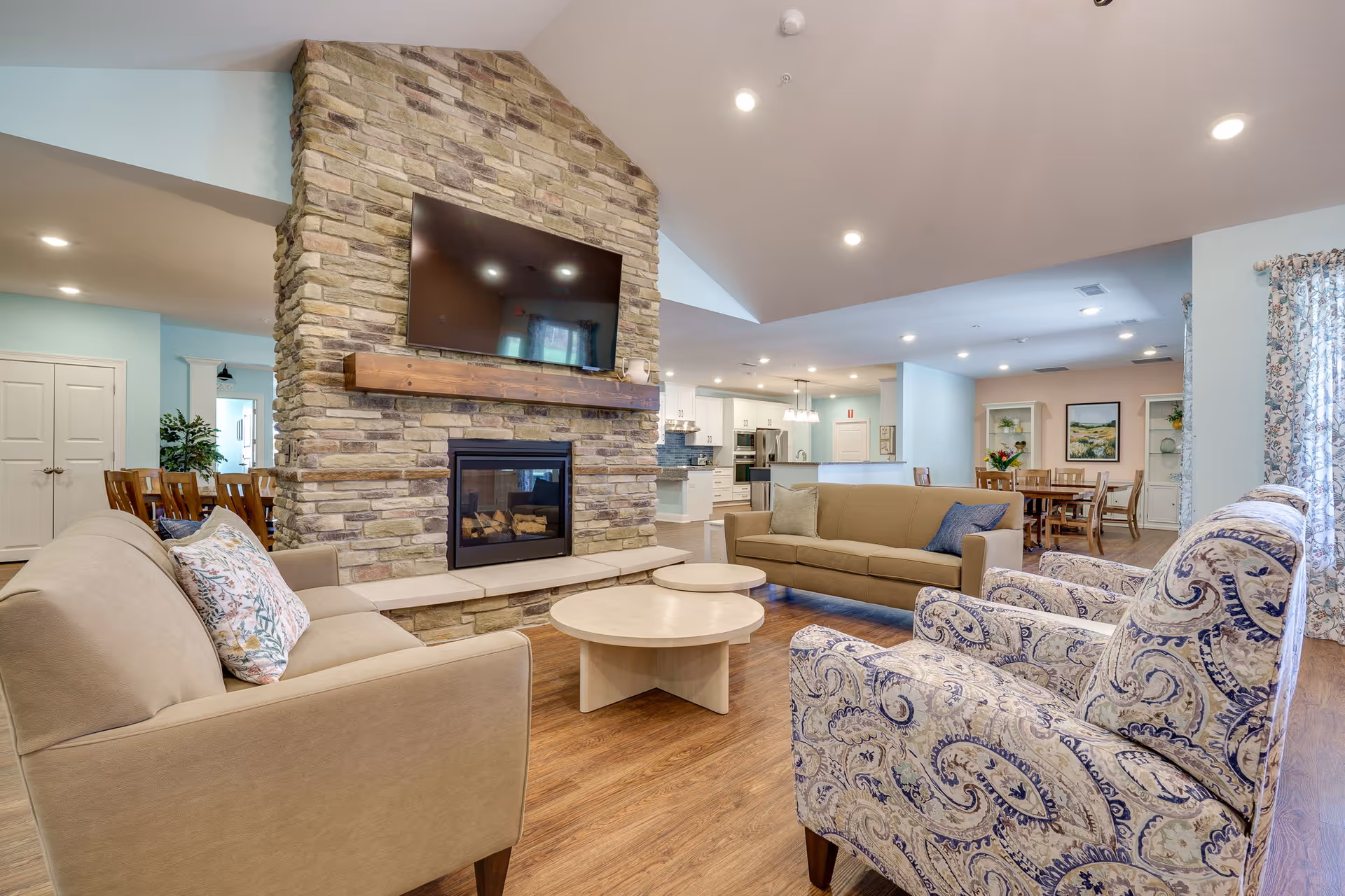 A spacious living room area in an assisted living facility featuring a stone fireplace with a mounted flat-screen TV above it. The room has two beige sofas, two patterned armchairs, and two round wooden coffee tables. The background shows a dining area with a wooden table and chairs, and a kitchen with white cabinetry and stainless steel appliances. The room has wooden flooring and light blue walls with floral curtains on the windows.
