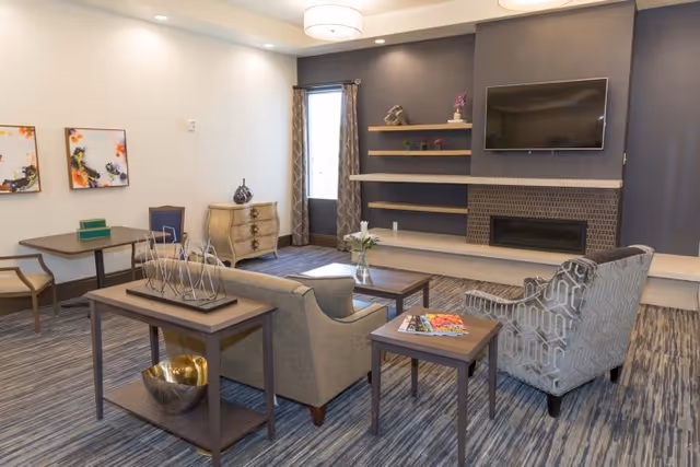 A modern communal living room with sofas, armchairs, a coffee table, shelving and a wall-mounted TV above a fireplace.