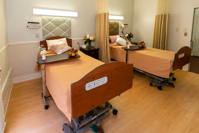 A healthcare facility room with two hospital beds separated by a curtain. Each bed has a peach-colored blanket, pillows, and a tray table with flowers and a fruit basket. The room has light-colored wooden flooring and soft lighting.