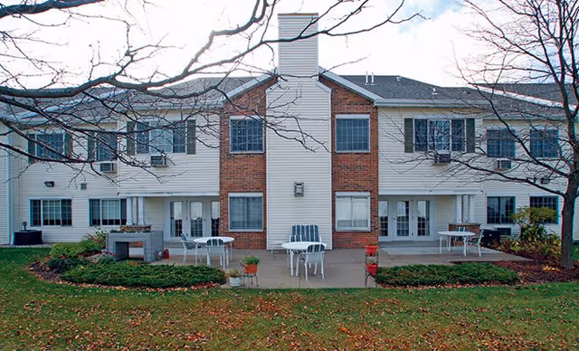 Exterior view of a two-story residential building with white siding and brick accents. The building has multiple windows and glass doors leading to small patios with white outdoor tables and chairs. Leafless trees and a grassy area with some fallen leaves are visible in the foreground.