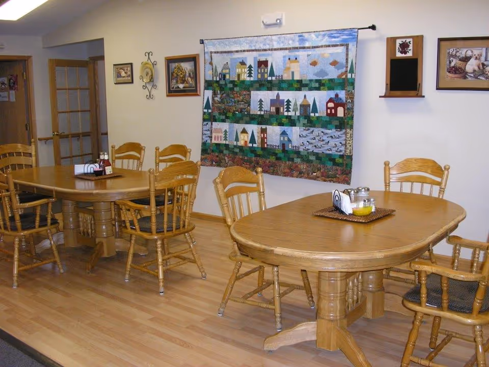 A dining area with two wooden tables, each surrounded by wooden chairs with cushions. On the tables are condiment holders with napkins, salt, pepper, and a small bowl. The walls are decorated with framed pictures and a colorful quilt hanging. The floor is light wood, and there is a door with glass panels in the background.
