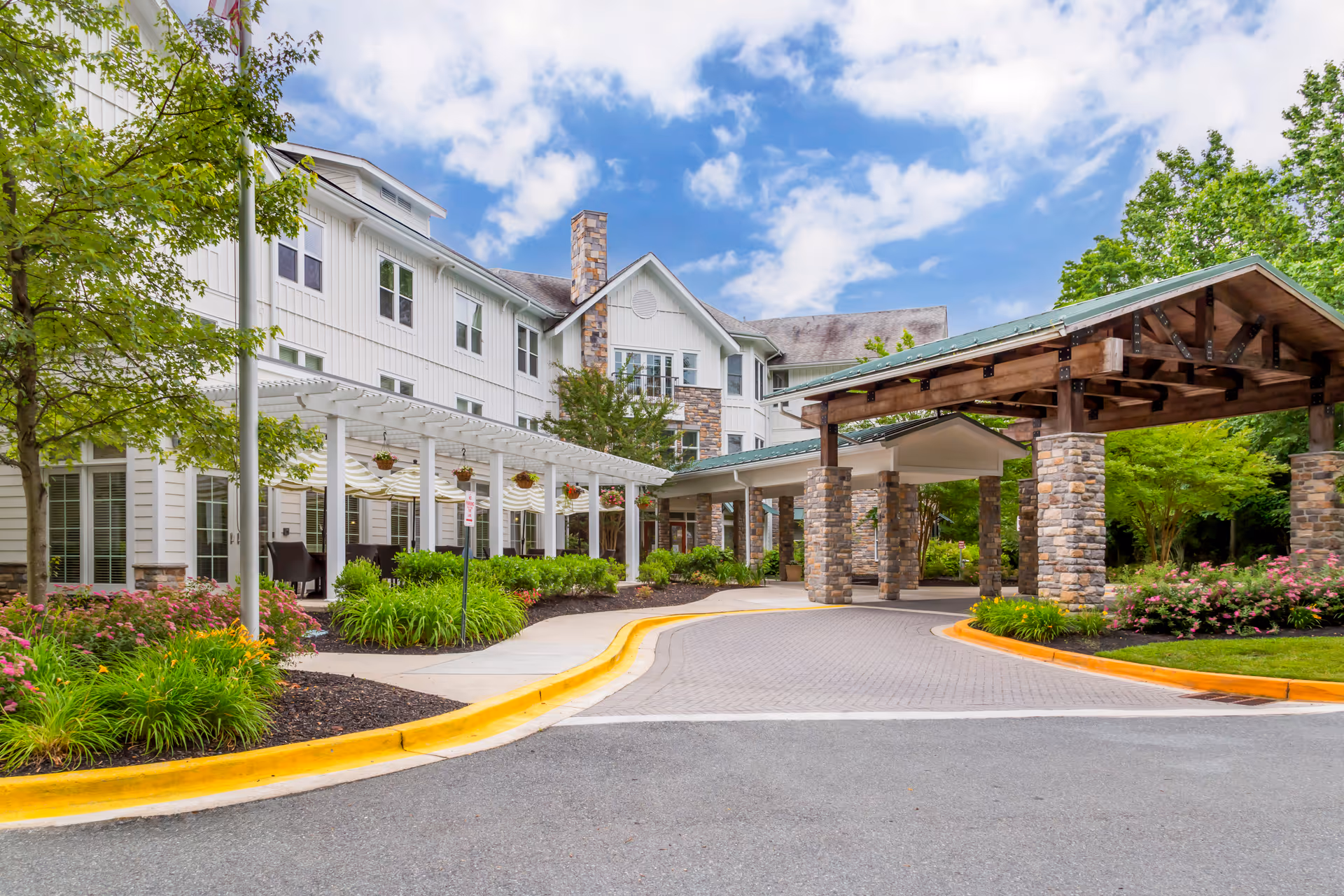 Exterior view of a senior living facility with a covered entrance supported by stone pillars, a white multi-story building with large windows, a pergola with hanging flower baskets, landscaped greenery, and a partly cloudy sky.
