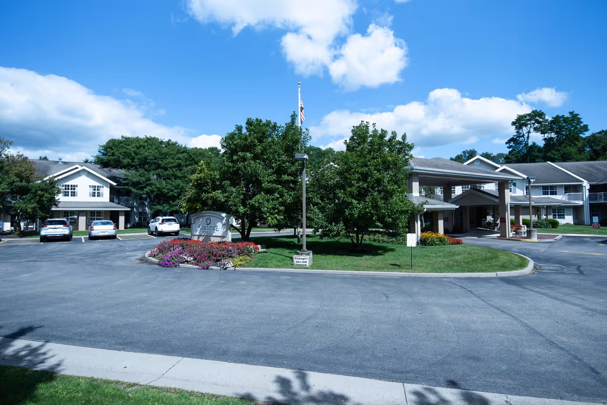 Exterior view of Solstice Senior Living at Fairport facility showing a driveway with parked cars, a landscaped area with trees and flowers, a flagpole, and the entrance to the building under a covered drop-off area.