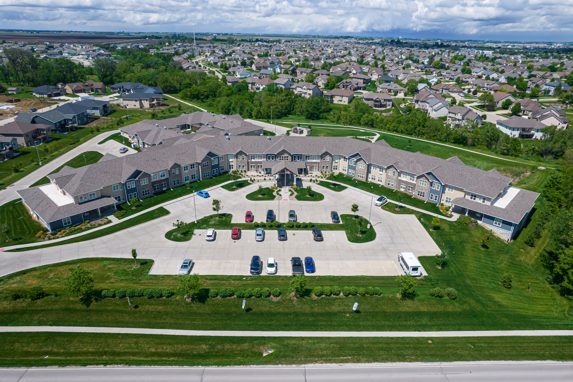 Aerial view of Rock Creek Senior Living facility showing a large, U-shaped building with a parking lot in front, surrounded by green lawns and trees. The facility is located in a suburban area with many houses visible in the background under a partly cloudy sky.