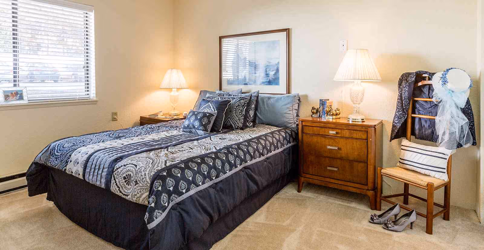 Cozy bedroom with a patterned bed, wooden nightstand with lamps, a window with blinds, and a chair holding clothing and shoes.