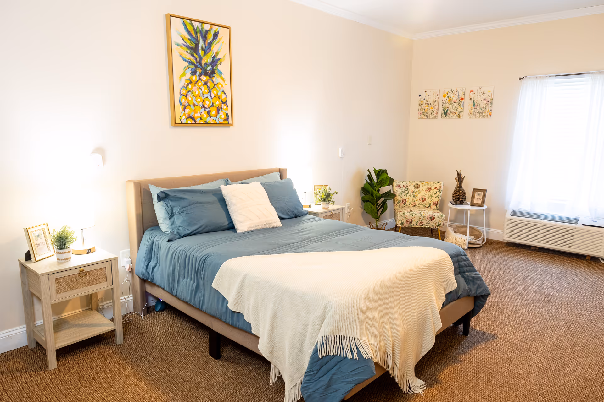 A bright and cozy bedroom with a neatly made bed featuring blue bedding and a cream throw blanket. There are two nightstands on either side of the bed, each with a small lamp and decorative items. A colorful pineapple painting hangs above the bed. In the corner, there is a floral-patterned armchair next to a small round table with a pineapple decoration and a framed photo. A large window with sheer white curtains lets in natural light, and a wall-mounted air conditioning unit is below the window.