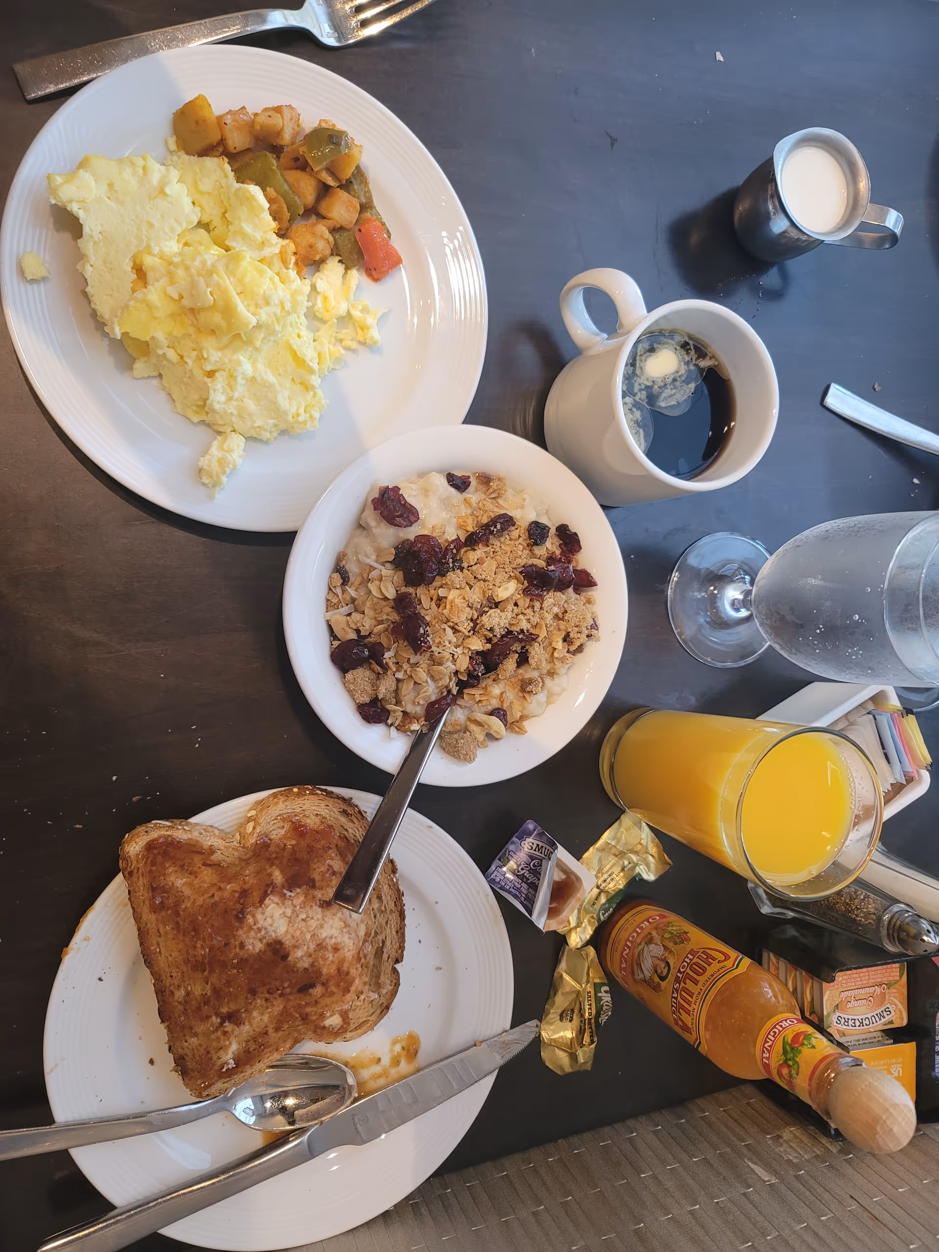 Breakfast spread on a dark table with scrambled eggs and potatoes, a bowl of granola/yogurt with dried cranberries, toast, coffee, and a glass of orange juice.
