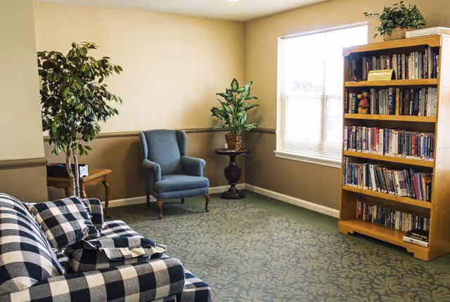 A cozy living room area with a checkered sofa, a blue armchair, two potted plants, a wooden bookshelf filled with books, and a window with blinds letting in natural light.