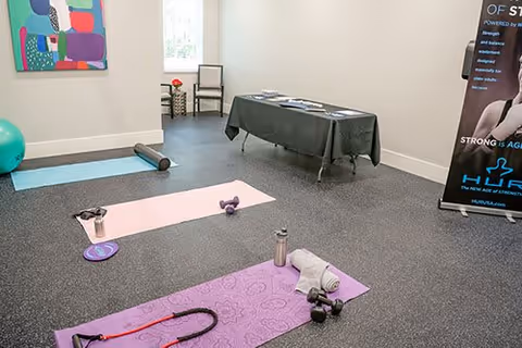 A small exercise room with yoga mats, dumbbells, a foam roller, a water bottle, and resistance bands on a rubber floor. There is a colorful abstract painting on the wall, a chair near a window, and a table covered with a black cloth in the background. A standing banner with fitness-related text is visible on the right side.