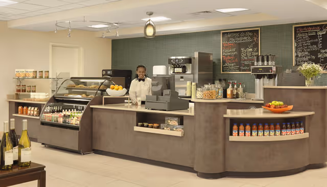 Indoor café-style service counter with a person behind the cash register, pastry display cases, bottled drinks, and menu chalkboards.