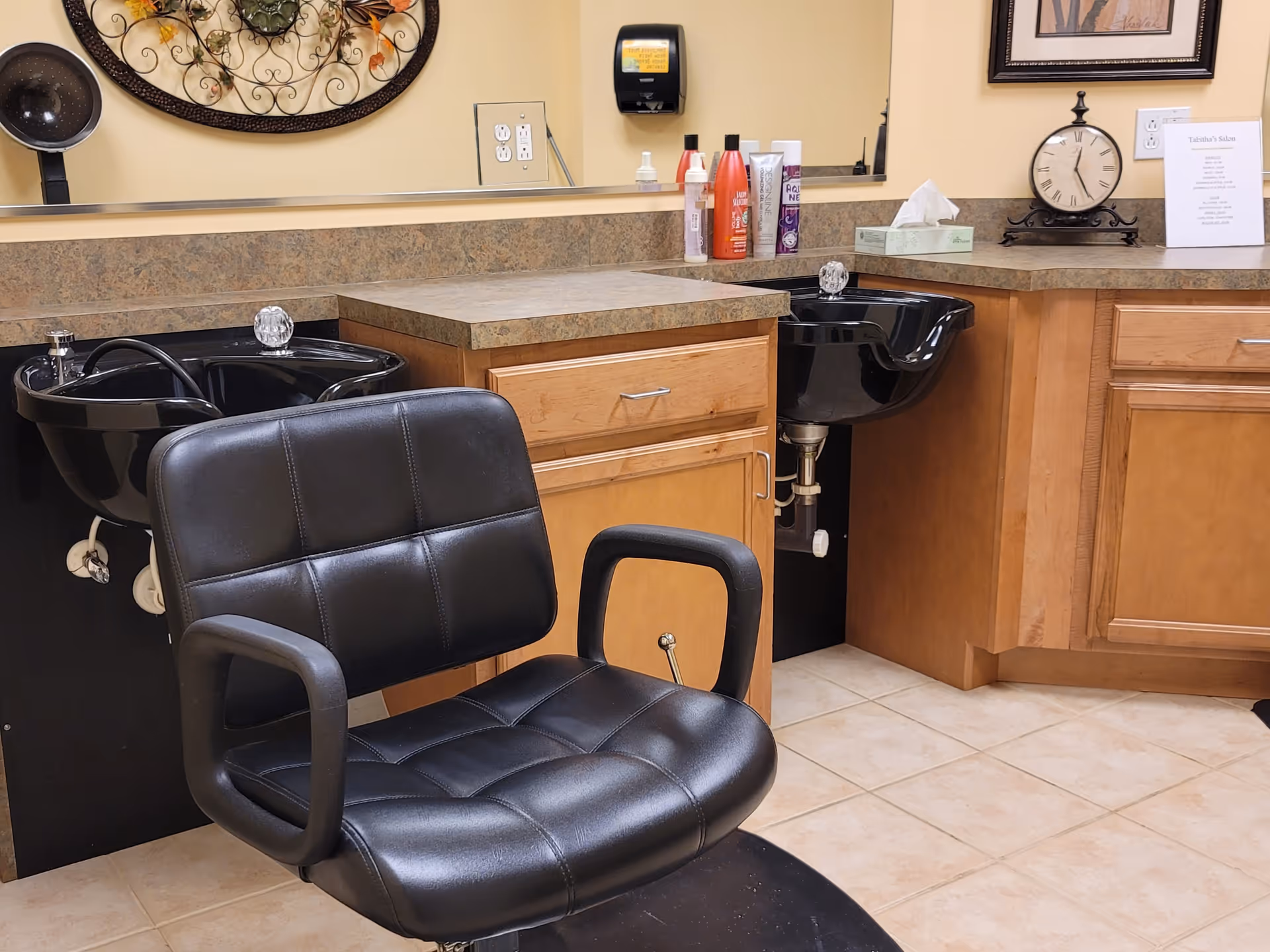 Interior view of a salon area in a senior living facility featuring a black salon chair in front of two black hair washing sinks. The countertop has various hair care products, a tissue box, a decorative clock, and a framed sign. The walls are painted beige, and there is a large mirror above the countertop.