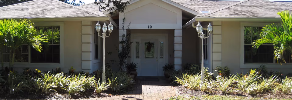 Front entrance of a single-story building with a brick walkway, ornamental lamp posts, palm plants, and double doors.
