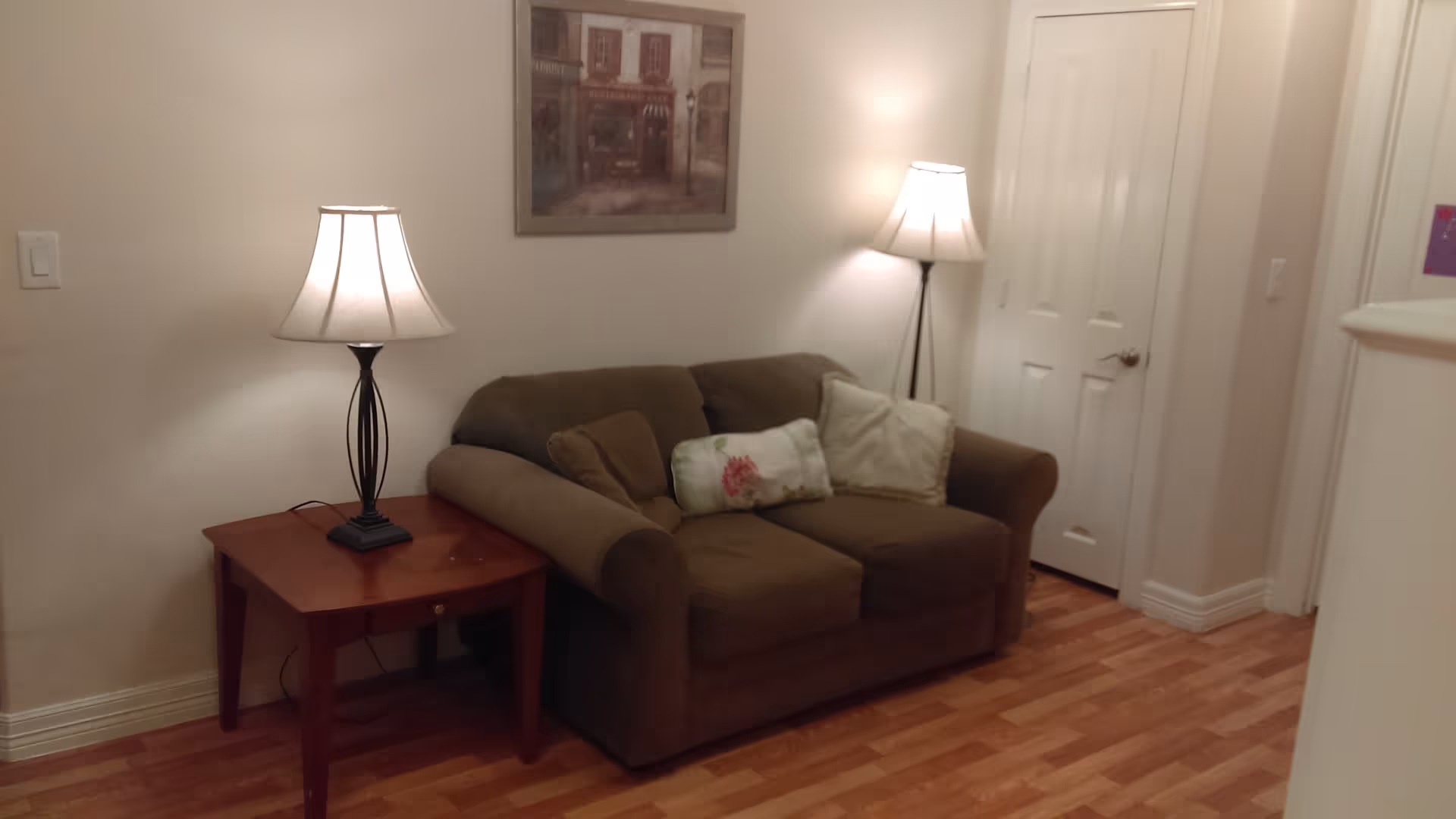 A cozy living room with a brown couch adorned with three pillows, two table lamps on either side of the couch, a wooden side table, a framed picture hanging on the wall, and a white door in the background. The floor is wooden and the walls are painted light beige.