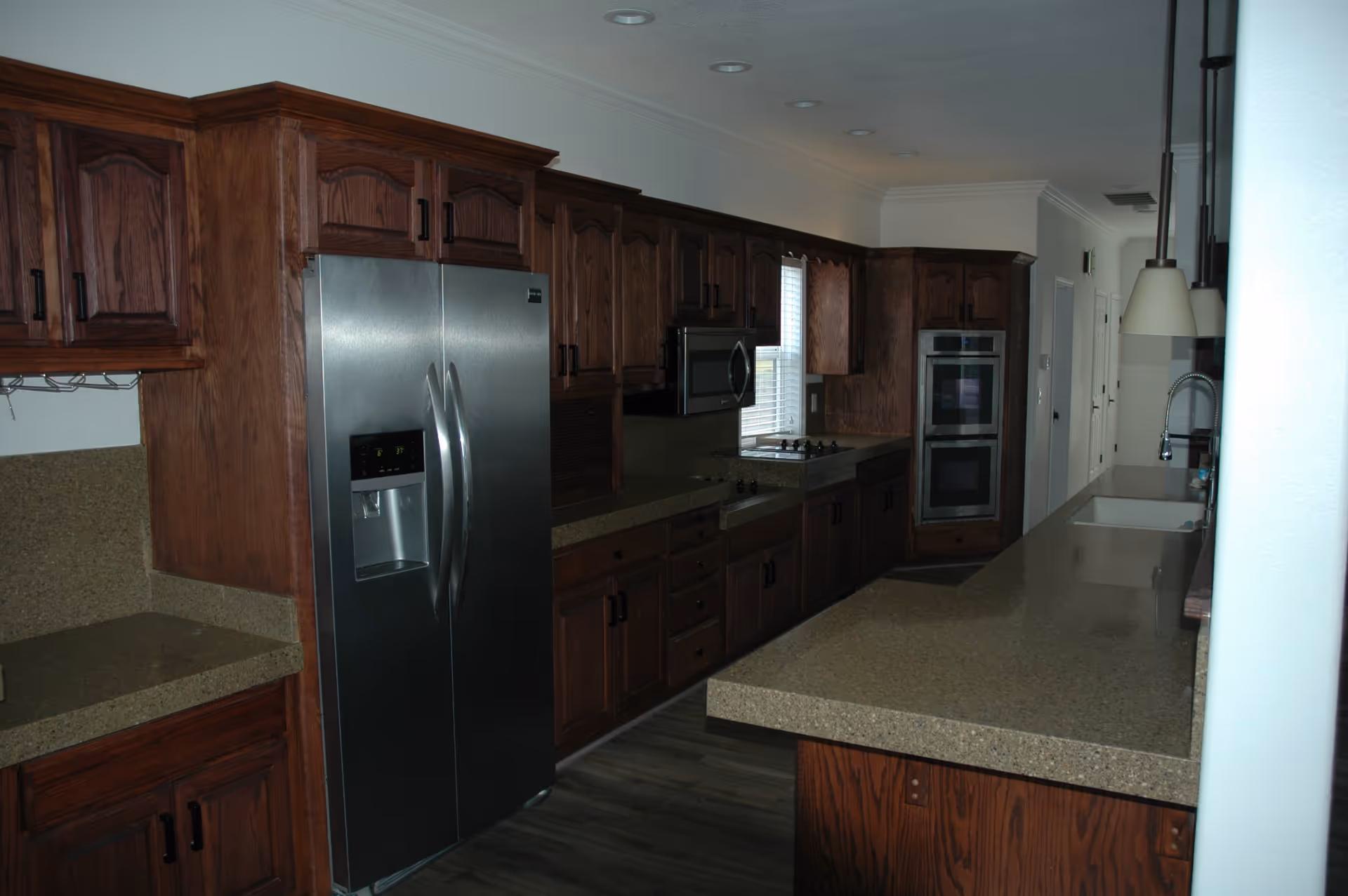 A long kitchen with dark wooden cabinets, a stainless steel refrigerator with water and ice dispenser, a built-in microwave, a stovetop, double ovens, and a long countertop with a sink and pendant lights hanging above.
