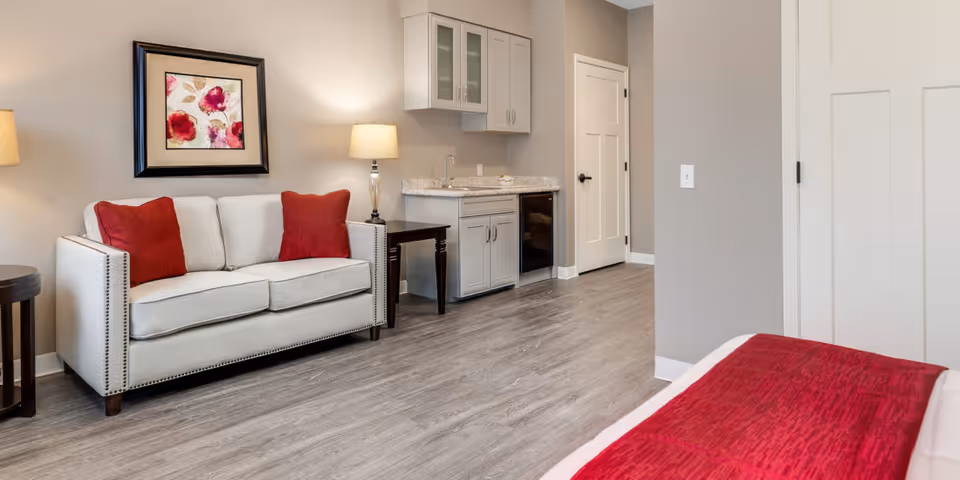 A cozy interior room featuring a light gray sofa with two red cushions, a side table with a lamp, a small kitchenette with a sink, cabinets, and a mini fridge, and part of a bed with a red blanket in the foreground. The walls are painted light gray and there is a framed floral artwork above the sofa.