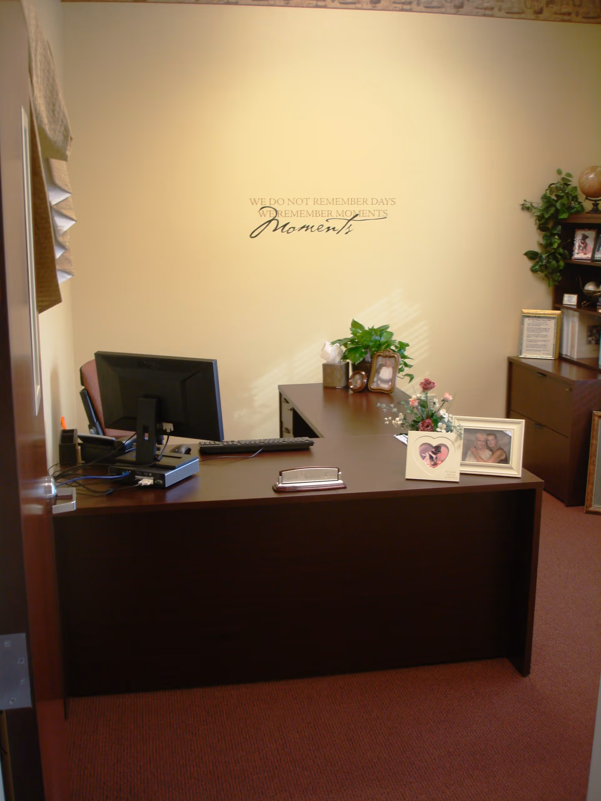 A tidy office reception with a dark wood desk, computer monitor, framed photos and plants against a beige wall with a decorative quote.