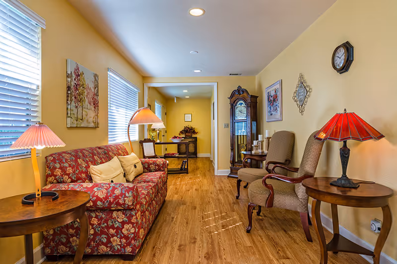 A cozy living room with yellow walls and wooden flooring featuring a floral patterned sofa with beige cushions, two wooden side tables each with a lamp, two upholstered armchairs, a grandfather clock, and wall decorations including a clock and framed artwork.