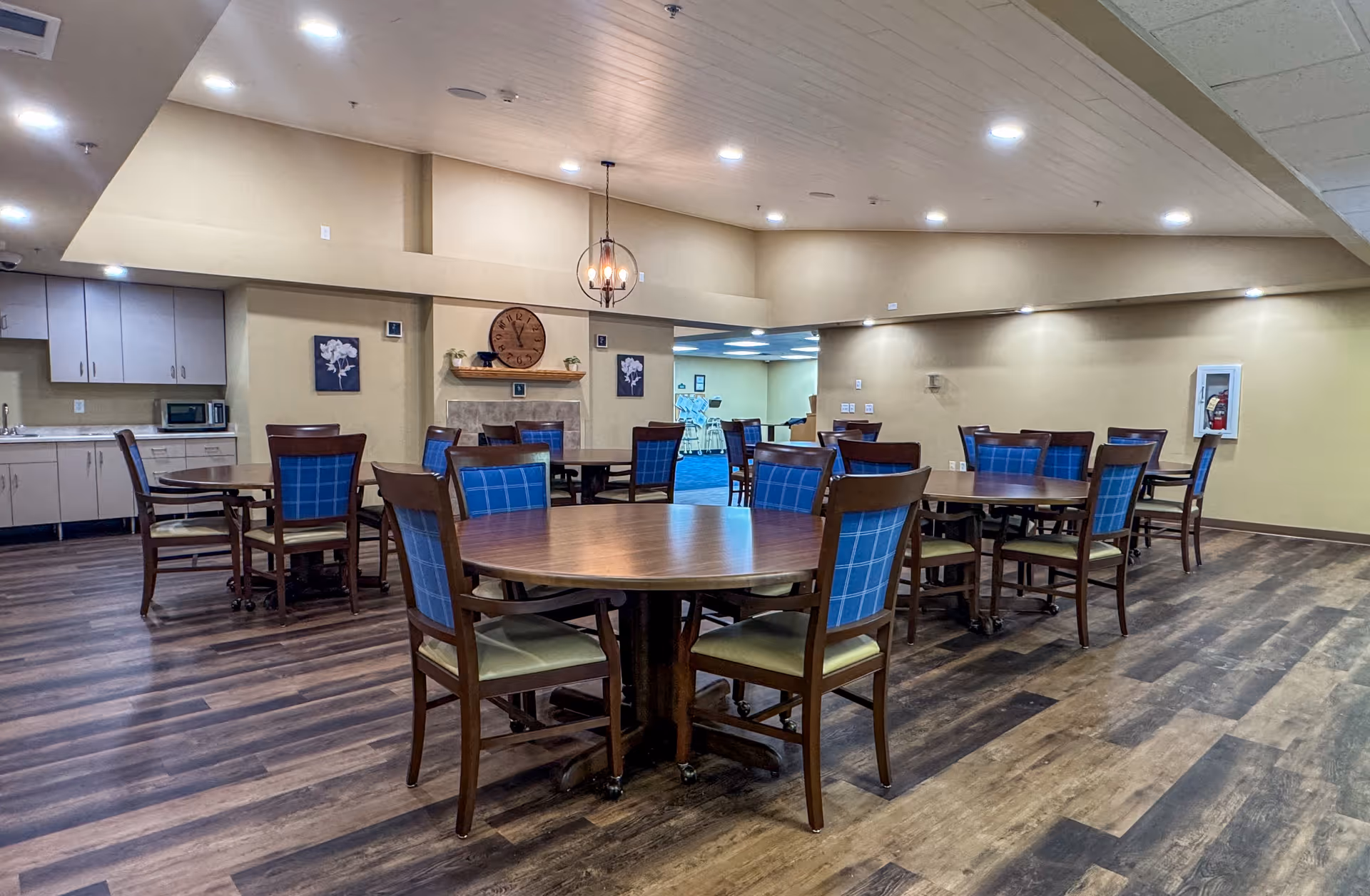 A spacious dining area with multiple round wooden tables and chairs with blue cushioned backs and beige seats. The room has wood-patterned flooring, beige walls, and a ceiling with recessed lighting. There is a small kitchen area with cabinets, a microwave, and a sink on the left side. A decorative clock and framed floral artwork are mounted on the wall above a fireplace. In the background, another room with blue carpeting and stacked chairs is visible.