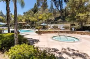 Outdoor pool and circular hot tub on a sunlit patio with lounge chairs, umbrellas, palm trees, and a hillside in the background.