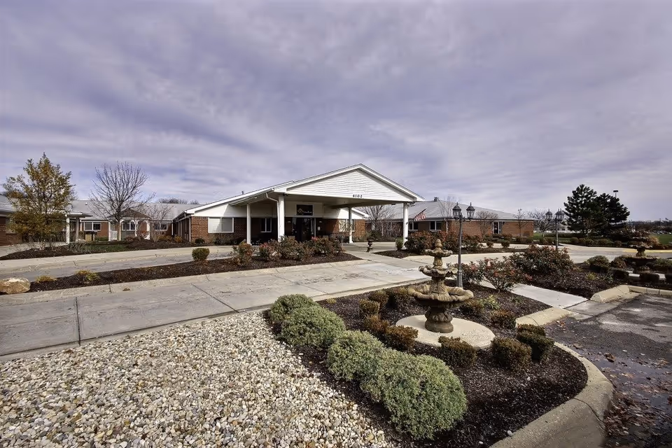 Exterior view of Eagle Creek Healthcare Center showing a single-story brick building with a covered entrance, surrounded by landscaped gardens with bushes, a stone fountain, and a paved driveway under a cloudy sky.