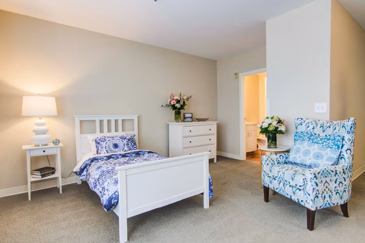 A bright and clean bedroom featuring a white single bed with blue and white patterned bedding, a white nightstand with a lamp and books, a white dresser with a vase of flowers and framed picture, and a blue patterned armchair with a matching pillow. An open doorway leads to a bathroom.