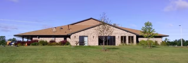 Single-story brick senior living building with a low brown roof set behind a large grassy lawn under a clear blue sky.