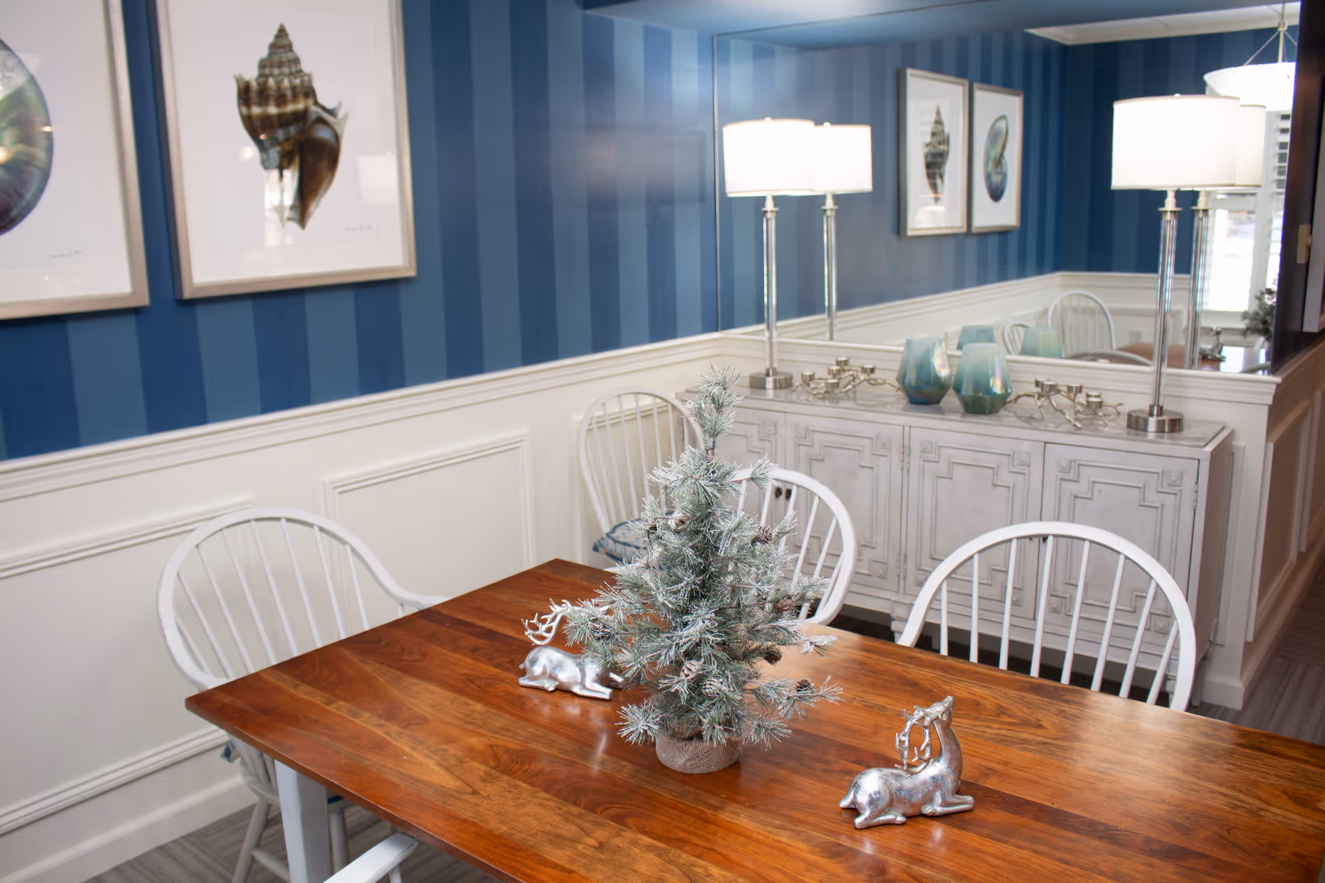 Dining room with a wooden table topped by a small frosted pine centerpiece and silver reindeer figurines, white spindle chairs, blue striped walls, and a mirrored sideboard with lamps.