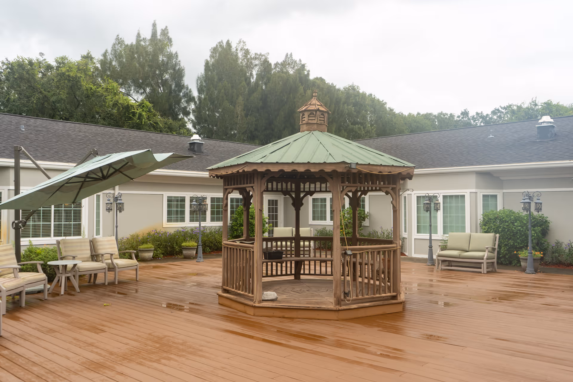 Outdoor patio area at The Colonnade at Northdale featuring a wooden gazebo with a green roof in the center, surrounded by cushioned chairs, a large umbrella, potted plants, and lamp posts. The patio is adjacent to a building with multiple windows and a gray roof, with trees visible in the background.