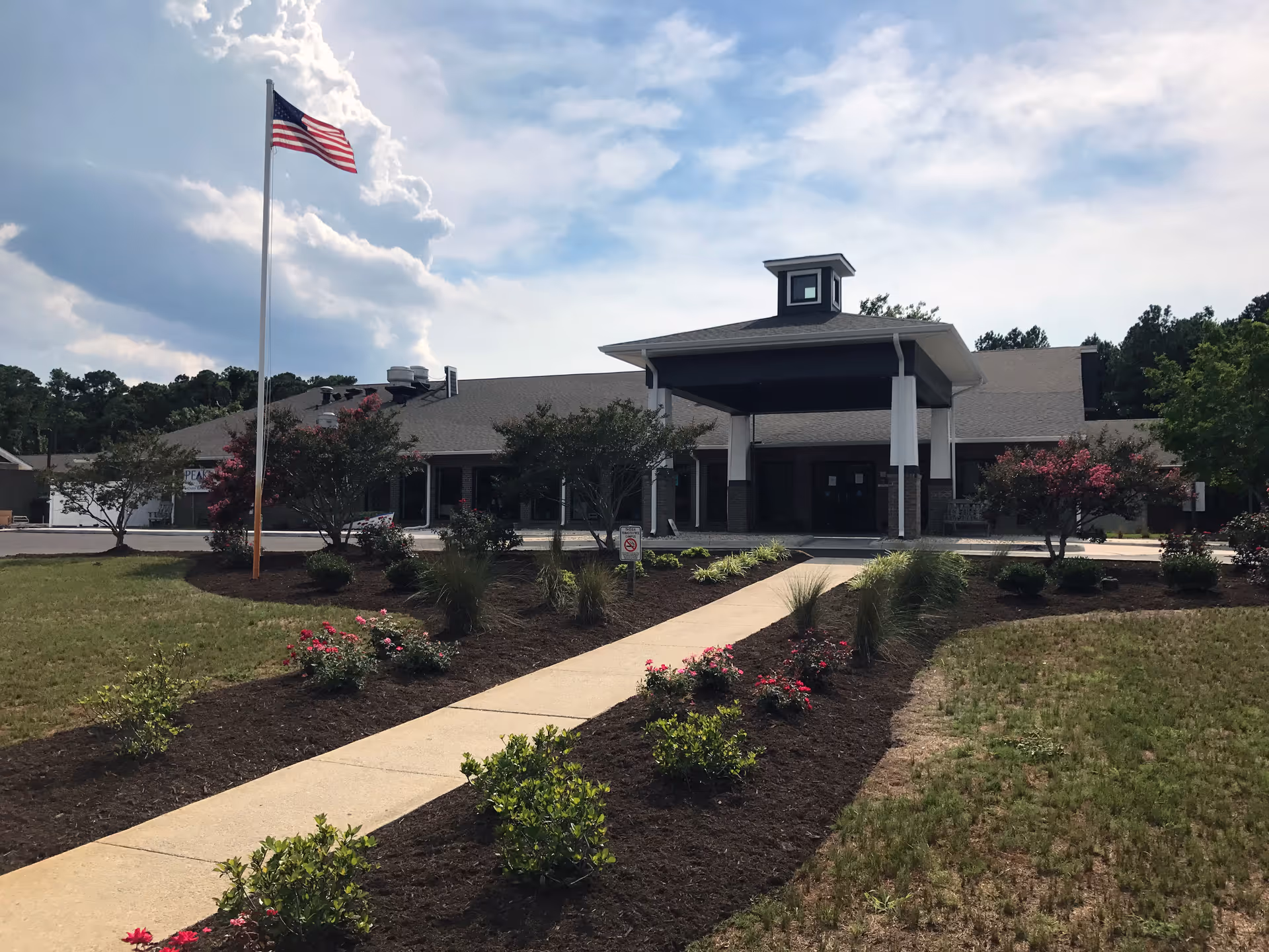 Front entrance of a single-story building with a covered porte-cochere, American flag, and landscaped walkway under a partly cloudy sky.