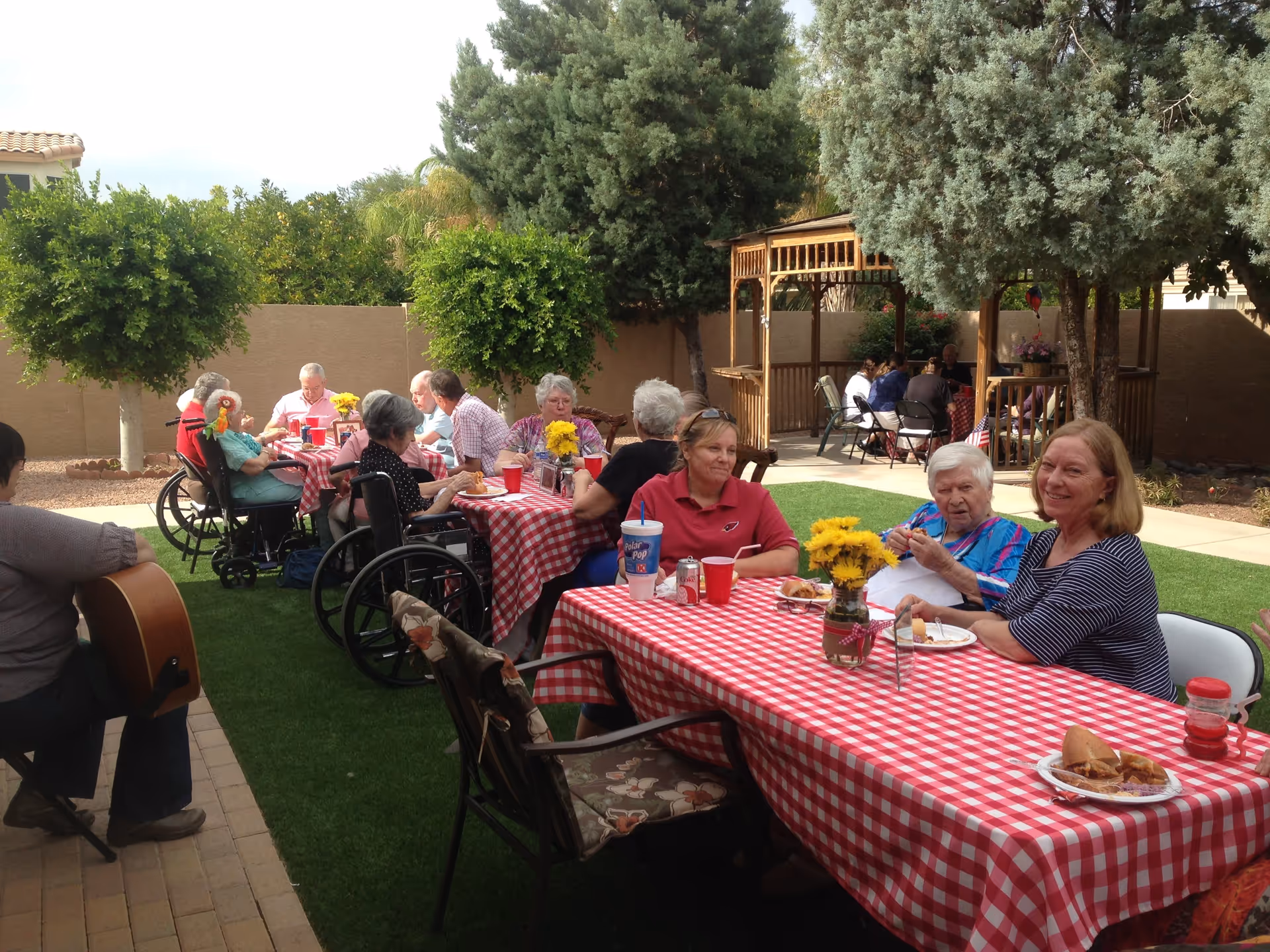 Older adults seated at red-checkered picnic tables in a landscaped courtyard with trees and a gazebo.