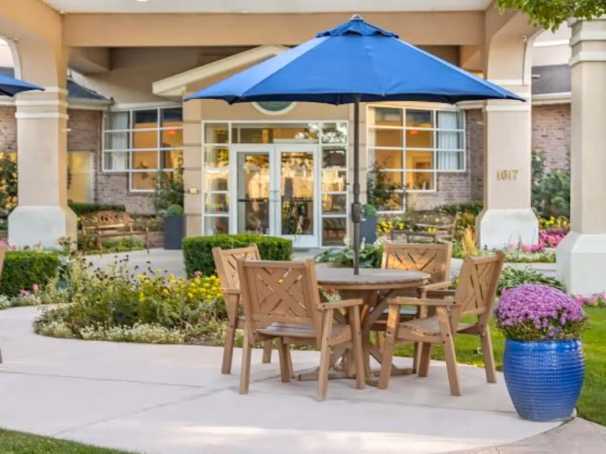 Outdoor patio area at Legacy Retirement Residence featuring a round wooden table with four matching chairs and a large blue umbrella. The patio is surrounded by well-maintained flower beds, shrubs, and a large blue planter with purple flowers. The building entrance with glass doors and windows is visible in the background.