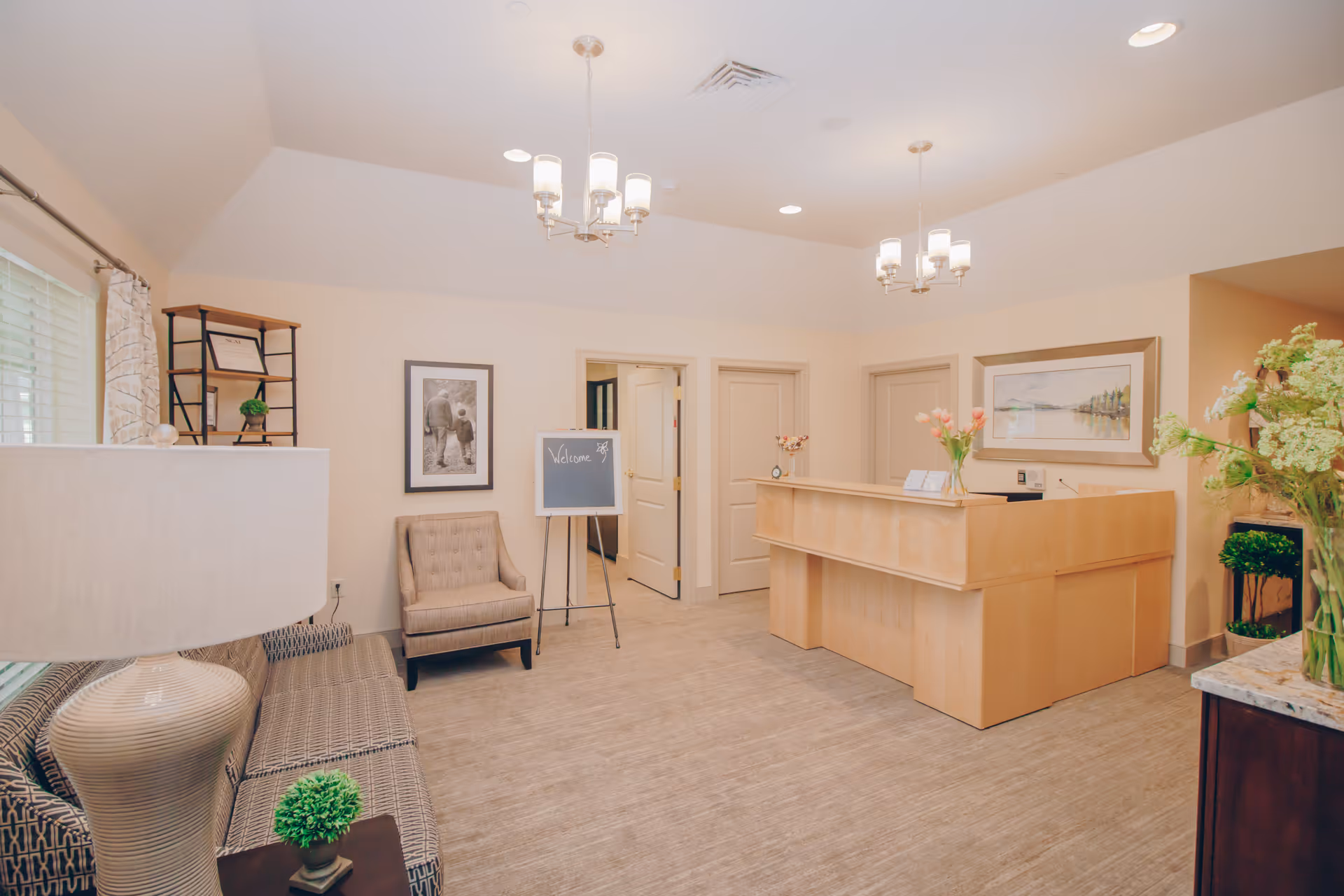 A bright and welcoming reception area in a senior living facility featuring a wooden reception desk with flowers, a seating area with a sofa and armchair, a lamp, framed artwork on the walls, and a small chalkboard sign that says 'Welcome'.