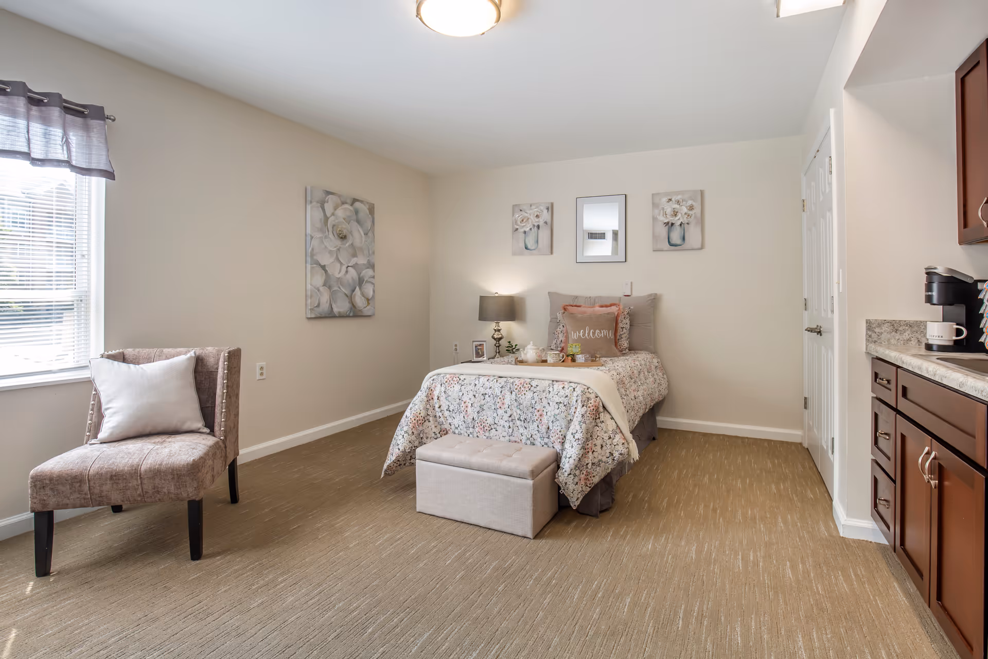 A cozy bedroom in a senior living facility featuring a single bed with floral bedding and a 'welcome' pillow. There is a small cushioned bench at the foot of the bed, a bedside table with a lamp and decorative items, a comfortable chair with a pillow near a window with blinds and a curtain, and three floral-themed wall art pieces above the bed. To the right, there is a kitchenette area with dark wood cabinets and a coffee maker.