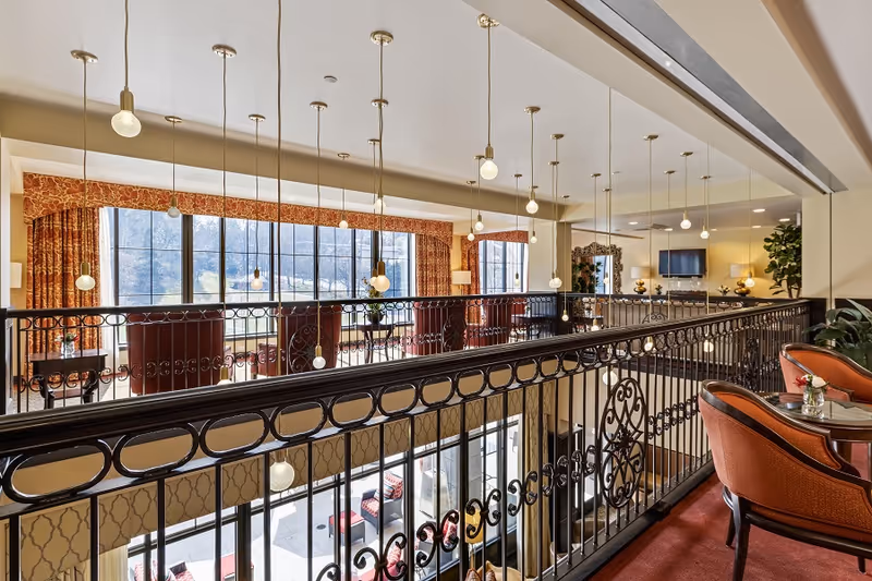 Upper-level lounge overlooking a lower seating area, with ornate iron railings, hanging pendant lights, and upholstered chairs by large windows.