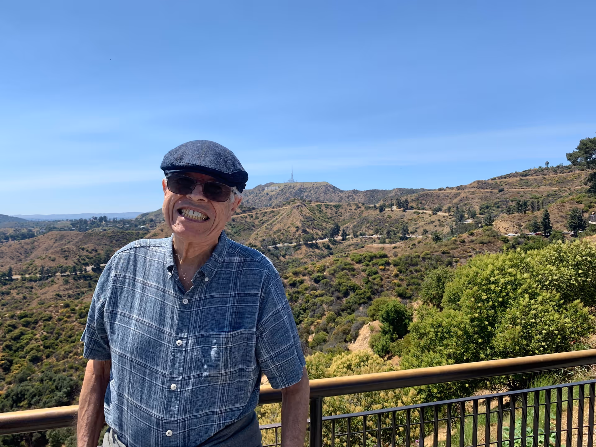 An elderly man wearing a blue plaid shirt, dark sunglasses, and a dark flat cap is smiling while standing outdoors on a balcony with a metal railing. Behind him is a scenic view of hills covered with greenery and a clear blue sky.