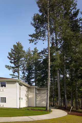 Exterior view of a two-story senior living facility building with beige siding, surrounded by tall trees and a curved concrete pathway leading to the entrance.