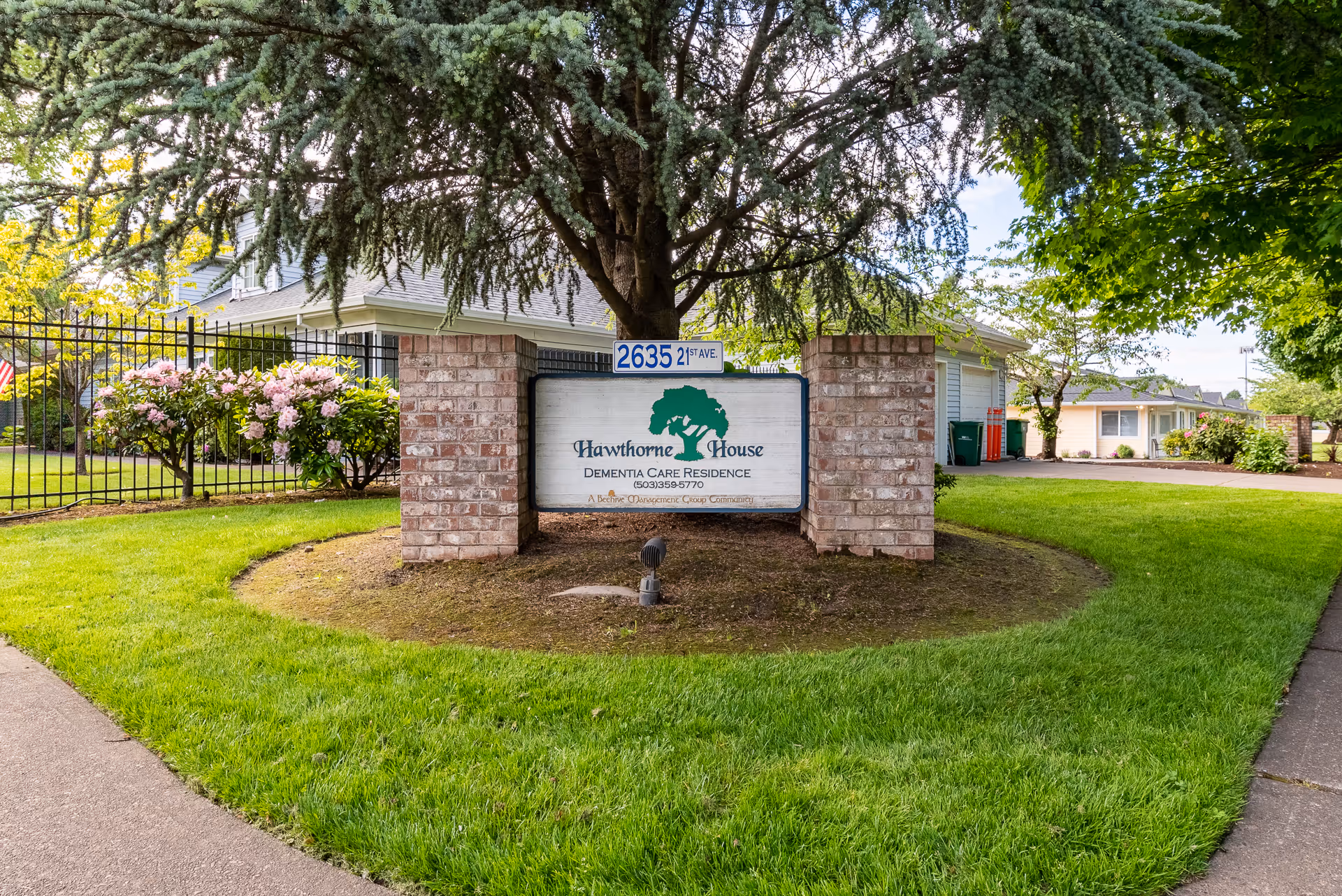 Outdoor view of the entrance sign for Hawthorne House Dementia Care Residence, featuring a brick structure with a white sign displaying the facility name and contact number, surrounded by green grass, trees, and flowering bushes.