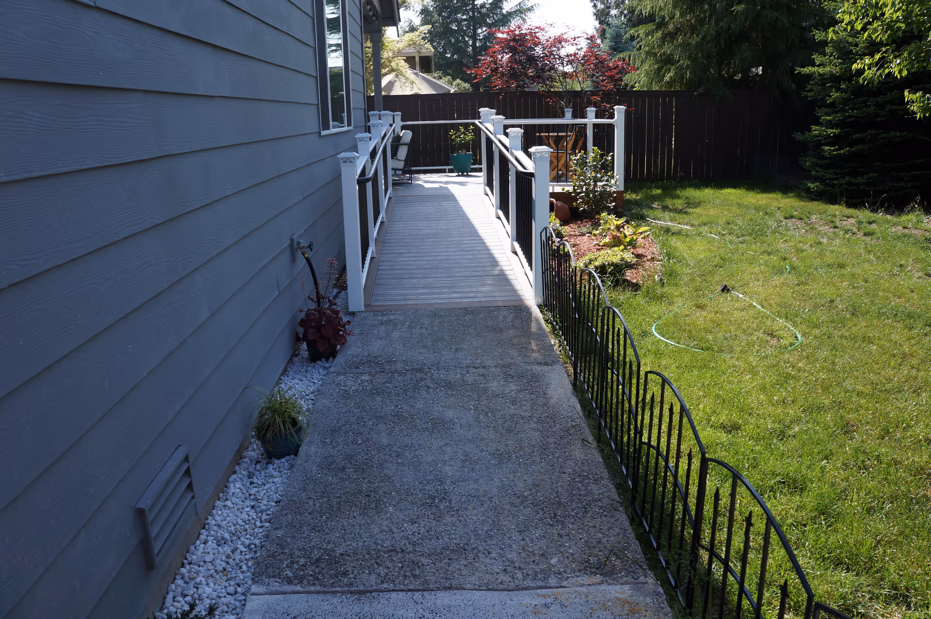 Outdoor view of a pathway alongside a gray building leading to a wooden ramp with white railings. On the right side, there is a small garden area with green grass, a few plants, and a black metal fence. A wooden fence and trees are visible in the background.