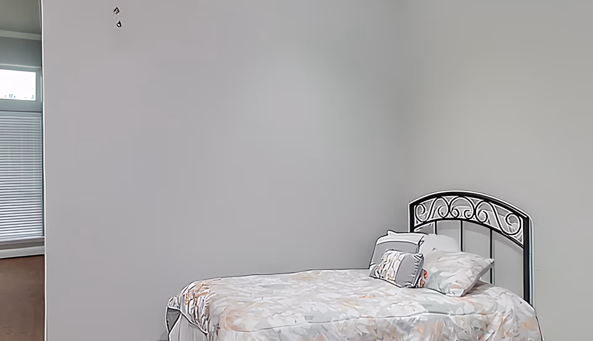 A simple bedroom with a metal headboard bed covered in a floral patterned bedspread and pillows. The walls are plain and light-colored, and there is a window with blinds partially visible on the left side.