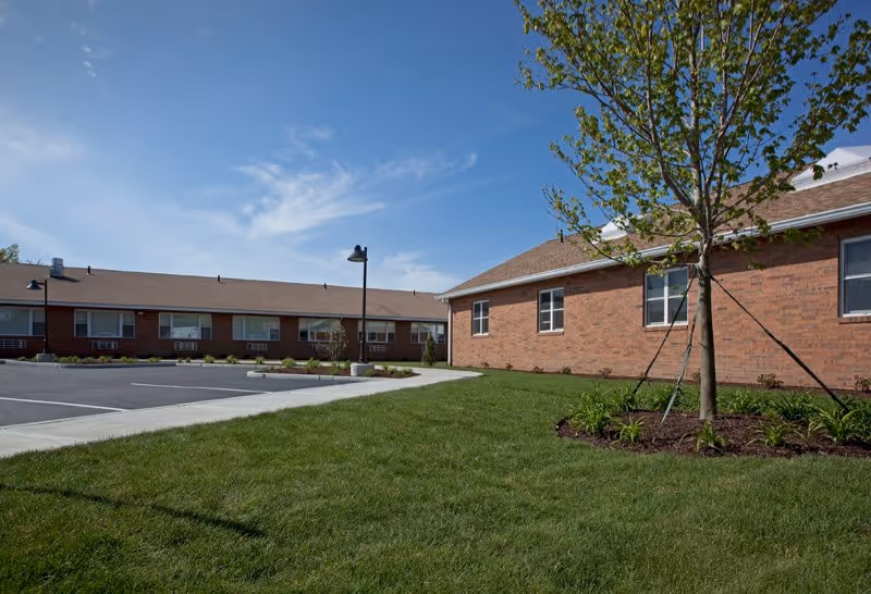 Brick single-story senior living buildings around a paved parking lot with green lawn and a young tree under a clear blue sky.