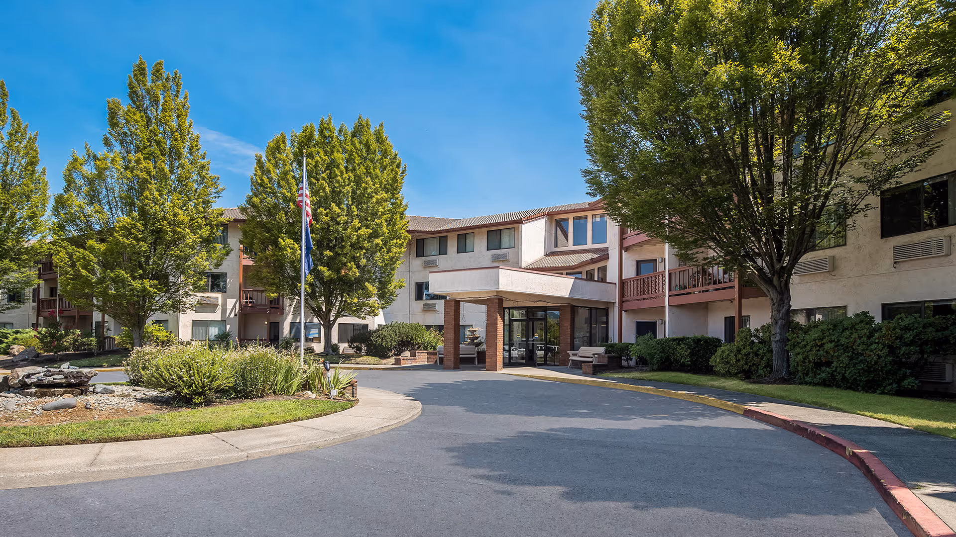 Exterior front entrance of a three-story senior living building with a covered drop-off, circular driveway, flagpoles, and landscaped trees and shrubs.
